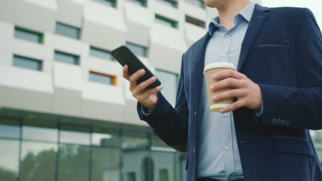 A-businessman-is-walking-near-the-business-center-with-a-cup-of-coffee-in-his-hand.-He-is-texting-on-a-smartphone.-He-is-wearing-a-suit-and-glasses.-Close-up-shooting.-4K