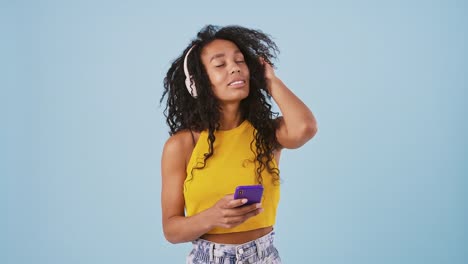 Afro-amerikanische-Frau-lächelt,-tanzt,-hört-die-Musik-über-Kopfhörer-und-schaut-auf-ihr-Smartphone.-Posieren-auf-blauem-Hintergrund