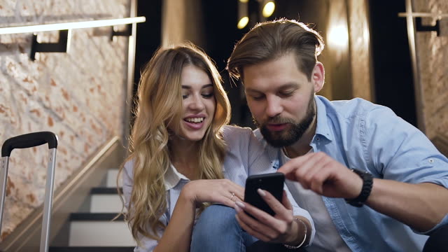 Close-view-of-happy-good-looking-blond-woman-and-beard-man-which-using-the-phone-while-sitting-on-steps-inside