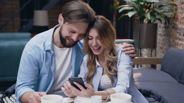 Cheerful-modern-stylish-young-couple-hugging-and-watching-at-woman's-phone-while-sitting-at-the-table-in-hotel-lobby