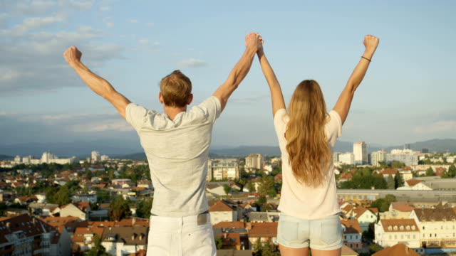 CLOSE-UP:-Young-couple-standing-on-rooftop-above-the-city,-raising-hands-in-sky