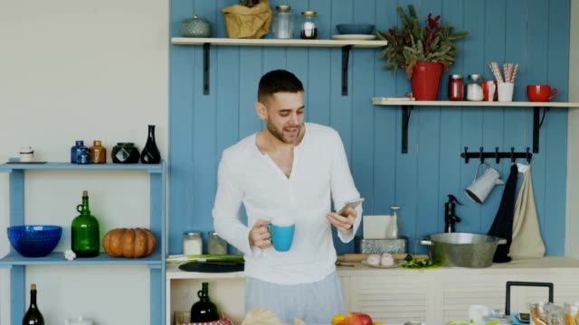 Slowmotion-of-Handsome-young-funny-man-dancing-and-singing-in-kitchen-while-surfing-social-media-on-his-smartphone-at-home-in-the-morning