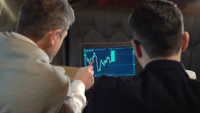 Two-colleagues-a-mature-man-and-young-man-discuss-trading-with-cryptocurrency-on-laptop-and-drink-coffee