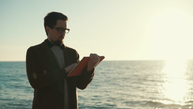 Man-is-standing-near-ocean,-holding-tablet-in-hands-and-touching-touchscreen