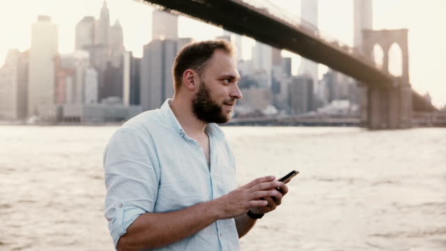 Feliz-caucásico-freelance-emprendedor-escribir-mensaje-en-smartphone,-sonriendo-cerca-de-Brooklyn-Bridge-Nueva-York-4K