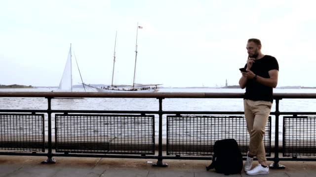 Young-handsome-man-standing-on-the-pier-near-the-fence-and-using-smartphone-near-the-water,-browsing-the-Internet
