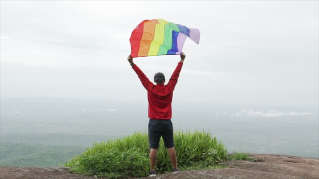 hombre-levantar-bandera-LGBTI-de-color-arco-iris-ondeando-en-duro-viento-en-el-mirador-de-alta-montaña