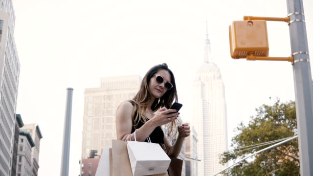 Trabajador-independiente-femenino-europeo-feliz-con-bolsas-de-la-compra-de-gafas-de-sol-sonriente,-usando-la-aplicación-para-smartphone-en-Nueva-York