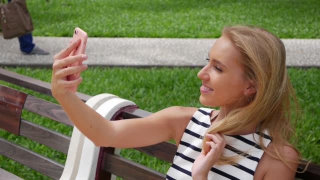 Young-beautiful-slim-woman-with-long-blonde-hair-in-black-and-white-dress-making-selfie-on-mobile-phone-over-background-the-park.