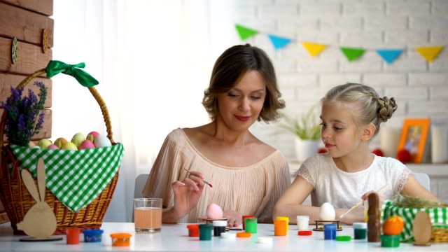 Madre-enseña-a-hija-colorear-huevos-de-Pascua,-preparando-para-vacaciones-familiares