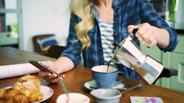 Mujer-vertiendo-café-mientras-revisa-el-teléfono-inteligente-en-la-mesa-del-desayuno