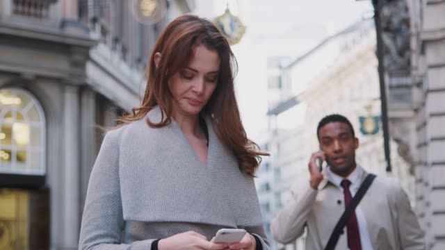 Millennial-white-woman-standing-on-the-street-using-smartphone,-young-black-businessman-talking-on-phone-walking-past,-close-up,-low-angle