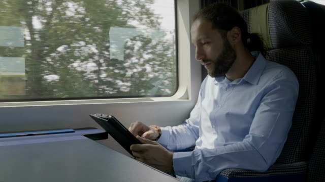 Focused-young-entrepreneur-swiping-and-scrolling-on-tablet-touchscreen-surfing-on-internet-next-to-window-in-moving-train