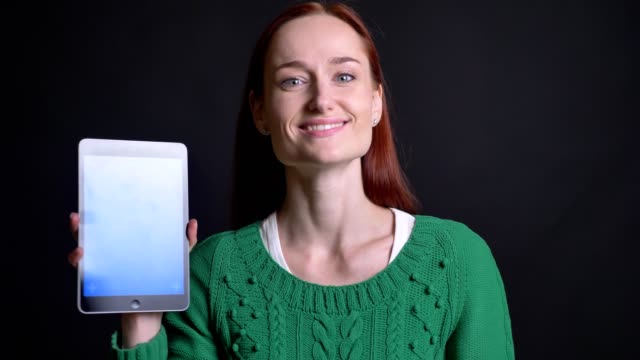 Closeup-portrait-of-attractive-caucasian-female-showing-the-screen-on-the-tablet-and-making-thump-up-looking-at-camera-and-smiling