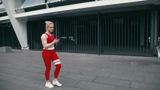 Plusgroße-blonde-gemischte-Rennracee-lächelnde-Frau-trägt-rote-Sportbekleidung-mit-Smartphone-im-Freien