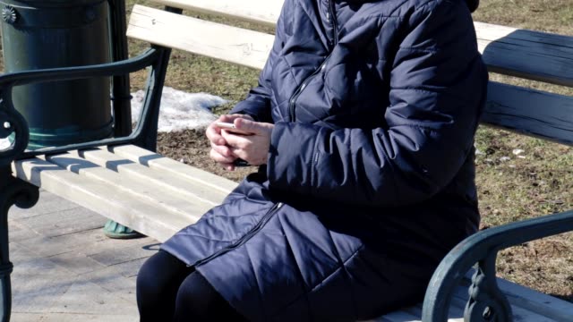 Eine-ältere-ältere-Frau,-die-auf-einer-Bank-im-Park-im-Freien-sitzt-und-mit-dem-Tapping-von-Smartphone-tippt-sie-ist-mit-dem-Smartphone,-um-mit-Menschen-auf-sozialen,-Textnachrichten-zu-verbinden-und-die-Suche-nach-Internet