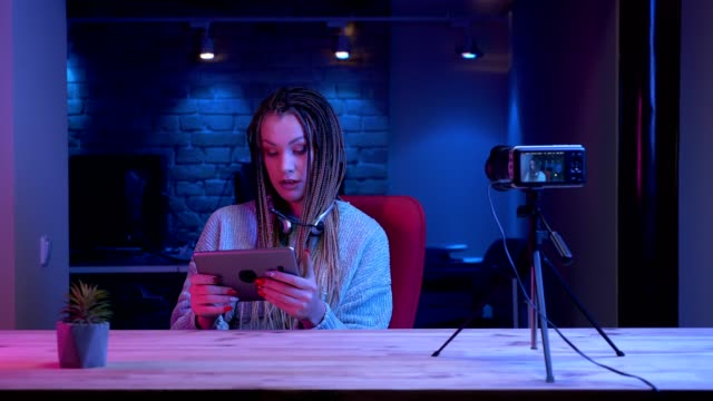 Closeup-shoot-of-young-female-blogger-with-dreadlocks-in-headphones-streaming-live-using-the-tablet-and-showing-green-chroma-screen-to-the-camera-with-the-neon-background