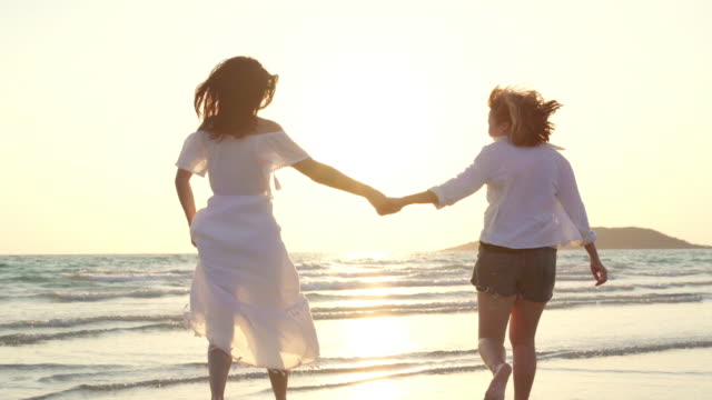 Junges-asiatisches-lesbisches-Paar-läuft-am-Strand.-Schöne-Freundinnen-freuen-sich,-sich-bei-Sonnenuntergang-am-Abend-am-Strand-am-Meer-zu-vergnügen.-Lifestyle-lesbisches-Ehepaar-reist-nach-Strandkonzept