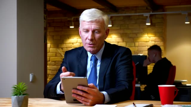 Closeup-shoot-of-old-caucasian-businessman-having-a-video-call-on-the-tablet-indoors-in-the-office-on-the-workplace