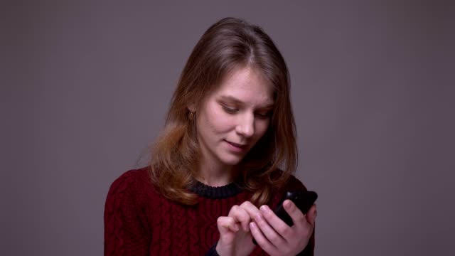 Young-female-student-working-with-smartphone-turns-to-camera-and-smiles-on-gray-background.