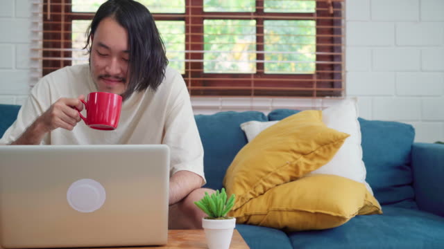 Freiberuflicher-asiatischer-Mann-arbeitet-zu-Hause,-männlich-kreativ-auf-Laptop-auf-dem-Sofa-trinken-Kaffee-im-Wohnzimmer.-Business-junge-Mann-Besitzer-Unternehmer,-spielen-Computer-am-Arbeitsplatz-bei-modernen-Hauskonzept.