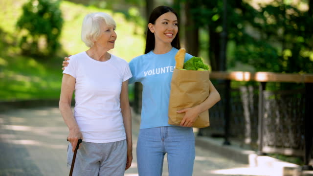 Social-volunteer-holding-grocery-bag-supporting-aged-woman-with-walking-stick