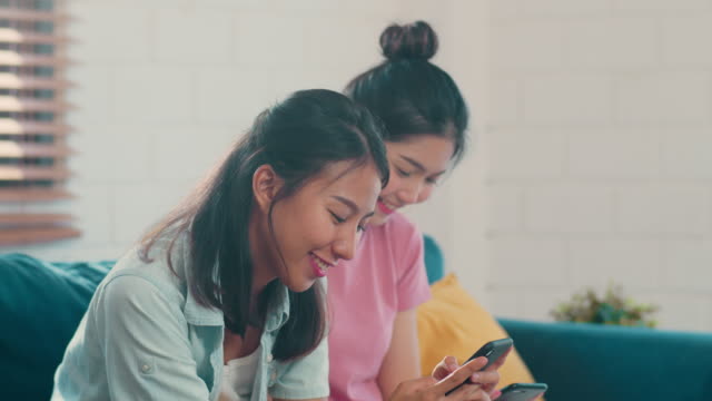 Junge-asiatische-Lesbische-Frauen-Paar-mit-Handy-überprüfen-Social-Media-im-Internet-zusammen,-während-liegen-Sofa-im-Wohnzimmer.