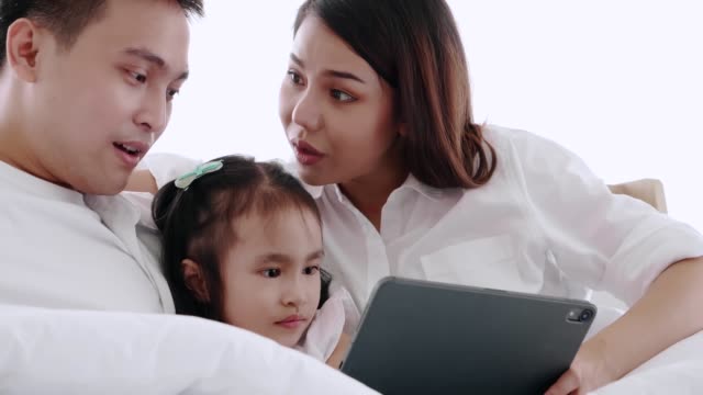 Asiatische-Familie-sitzt-auf-dem-Bett-lachend-und-suchen-in-digitalen-Tablet,-während-im-Schlafzimmer-liegen