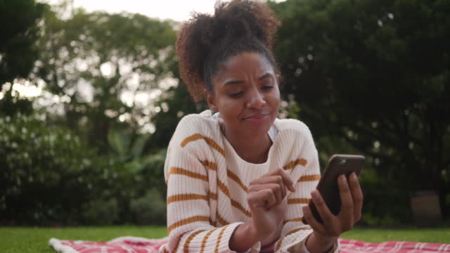 Porträt-einer-jungen-Afrikanerin,-die-im-Park-liegt-und-SMS-Nachrichten-auf-dem-Smartphone-genießt