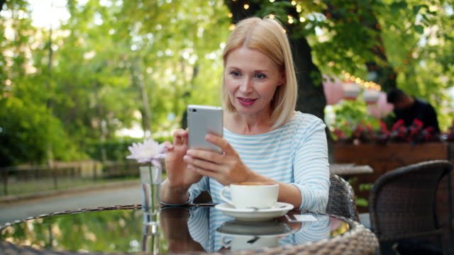 Cámara-lenta-de-la-mujer-feliz-usando-la-pantalla-táctil-del-teléfono-inteligente-en-el-café-exterior