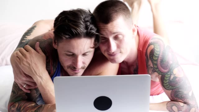 Gay-couple-in-bed-using-laptop-computer.-Hugging.