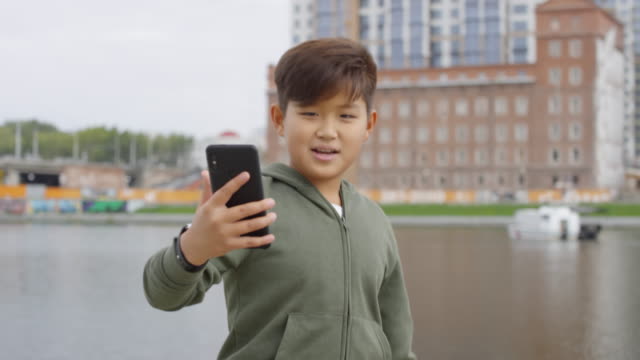 Asiático-Niño-Caminando-al-aire-libre-y-Hablando-a-través-de-Videollamada-en-Smartphone
