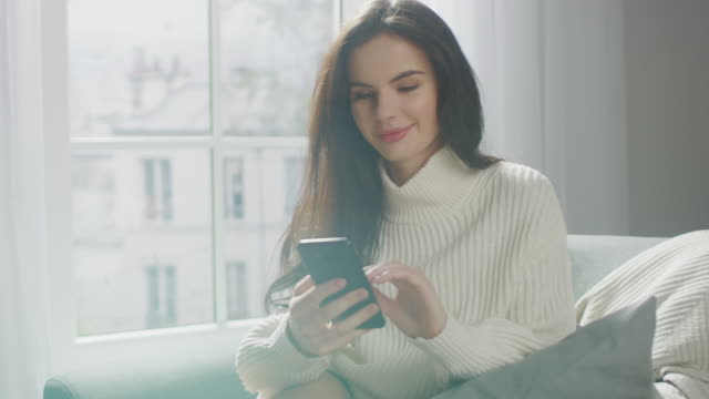 Beautiful-Young-Woman-Using-Smartphone-Happily,-while-Sitting-on-Chair.-Sensual-Girl-Wearing-Knitted-Sweater,-Surfs-Internet,-Posts-On-Social-Media,-Sharing-Picture-while-Relaxing-in-Cozy-Apartment