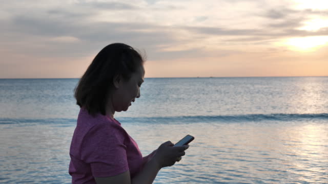 Nahaufnahme-Seitenblick-asiatische-Frauen-entspannen-zu-Fuß-am-Strand-und-verwenden-ein-Handy-am-Abend,-Sonnenuntergang.-Urlaubs--und-Reisekonzept.-Zeitlupe
