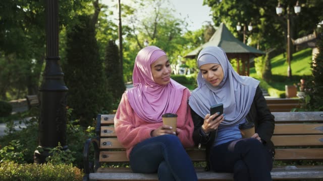 Two-muslim-females-in-casual-clothes-and-hijabs.-They-smiling,-enjoying-coffee-and-looking-at-screen-of-smartphone.-Sitting-on-bench-in-park.-Сlose-up