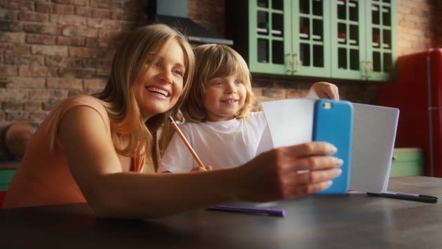 Lächelnde-Mutter-und-Kind-genießen-Online-Videoanruf-auf-dem-Smartphone,-während-Hausaufgaben-in-der-Schule-Copybook.-Sie-sitzen-in-der-Küche-am-Tisch.-Nahaufnahme