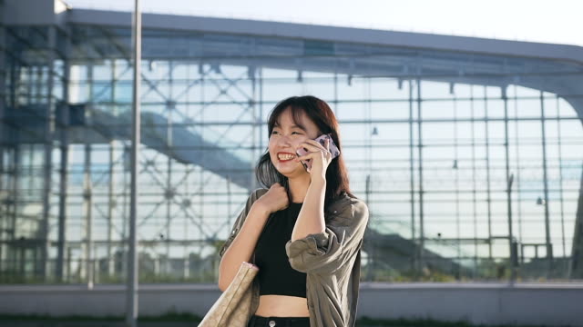 Pretty-cheery-young-asian-woman-with-dark-hair-talking-on-phone-and-sincerelly-smiling-near-airport-building-in-day-time