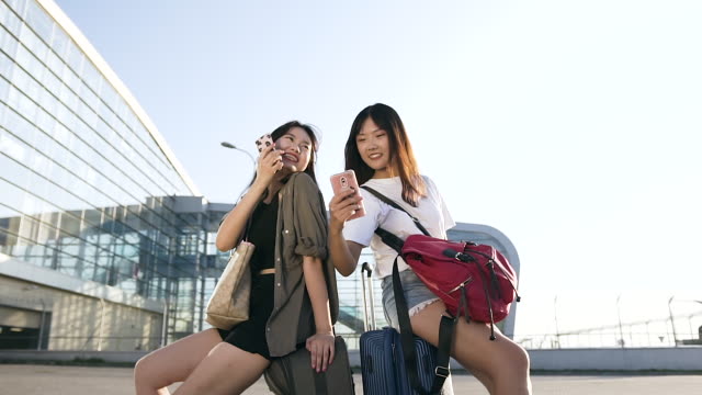 Felices-novias-asiáticas-de-25-años-de-edad-apuestos-sentados-en-sus-maletas-y-usando-teléfonos-inteligentes-cerca-de-la-moderna-terminal-del-aeropuerto