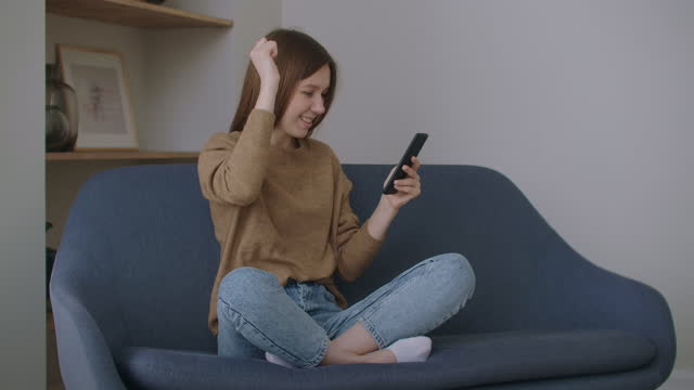 Young-woman-sitting-at-desk-at-home-office-distracted-from-work-holding-smart-phone-using-social-networking-website-chatting-with-friend-remotely-using-modern-device-and-internet-connection