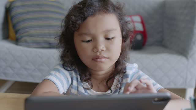 Niña-asiática-usando-tableta-digital,-jugando,-mientras-está-sentada-en-la-sala-de-estar-en-casa
