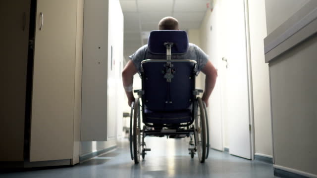 Vista-posterior-del-hombre-discapacitado-empuja-a-sí-mismo-en-silla-de-ruedas-por-el-pasillo-del-hospital