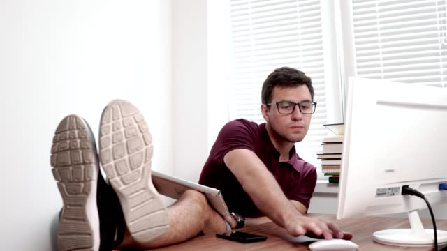 Joven-trabajador-en-gafas-sujeta-el-teclado-sobre-las-piernas-escribiendo-y-mirando-a-la-pantalla-de-la-computadora-en-la-oficina-con-sus-piernas-sobre-la-mesa.-Trabajador-cansado-ocupado-en-la-oficina-moderna.-4k