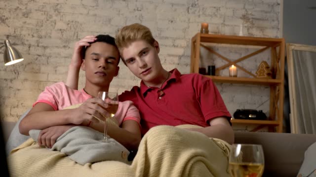 Multinational-gay-couple-sitting-on-couch-covered-with-a-warm-blanket,-watch-TV,-use-the-remote-control,-look-at-the-camera.-Homeliness,-romantic-evening,-background,-hugs,-happy-LGBT-family-concept.-60-fps
