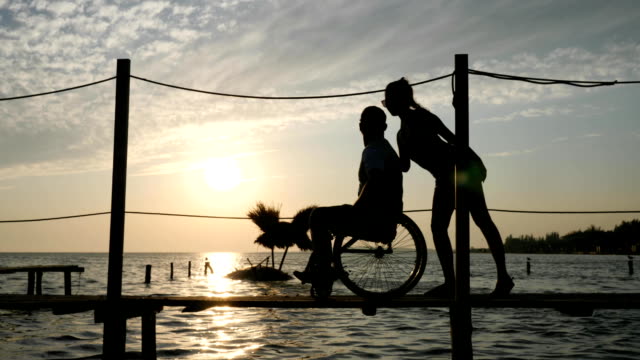 Disabled-person-living-a-full-life,-young-women-with-disabled-male-in-a-wheelchair-on-the-pier-at-waterfront,-background-heaven-in-orange-Sun-rays