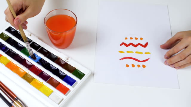 Nicht-erkennbare-Frau-malt-eine-Skizze-mit-hellen-Farben-auf-Papier-für-eine-Osterkarte.-Auf-dem-Tisch-ist-eine-Palette-mit-Farben-und-ein-Glas-Wasser.