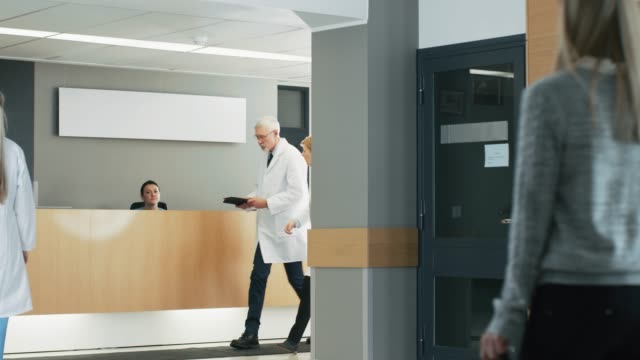 En-el-Hospital,-los-médicos-ocupados-hablan,-utilizando-Tablet-PC-mientras-camina-por-el-edificio.-En-el-paciente-antecedentes-habla-con-la-recepcionista.-Nuevo-centro-médico-totalmente-funcional-moderna.