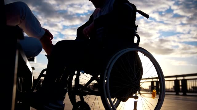 Silhouette-of-young-woman-with-disabled-man-in-a-wheelchair-talking-at-sunset-outdoors