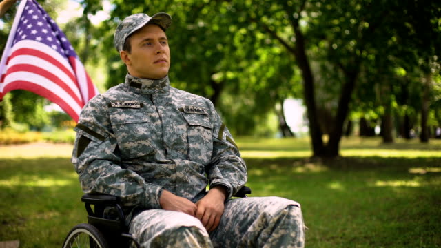 Veterano-saludando,-sentado-en-silla-de-ruedas,-ondeando-bandera-detrás,-día-de-la-independencia