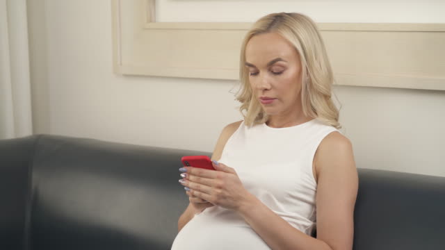Eine-schöne-schwangere-Blondine-auf-dem-Sofa-sitzen,-mit-Blick-auf-das-Telefon-ernst-und-etwas-eingeben