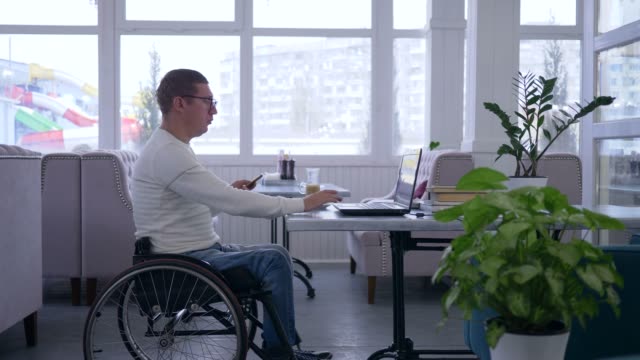 remote-business-management,-handicapped-man-in-wheelchair-uses-a-cellphone-sitting-at-table-with-laptop-in-cafe-on-the-background-of-large-window